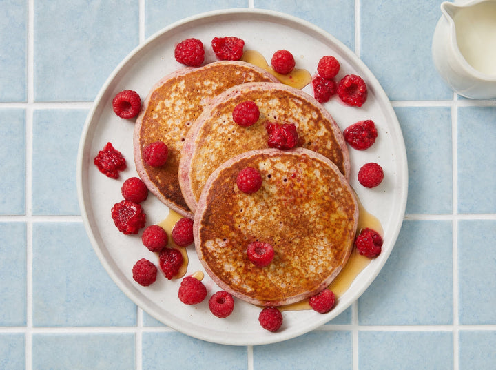 Vier Pancakes mit rosa Rand auf einem Teller, mit frischen Himbeeren und Ahornsirup