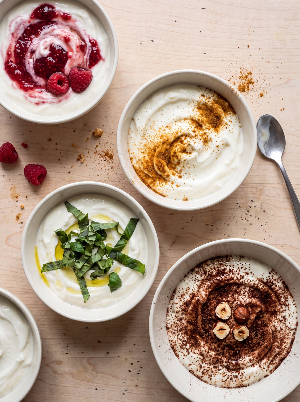 Vier Schalen Naturjoghurt auf hellem Holz, jede anders: mit Himbeere, mit Gewürz, mit Basilikum und Öl, mit Kakao und Haselnuss