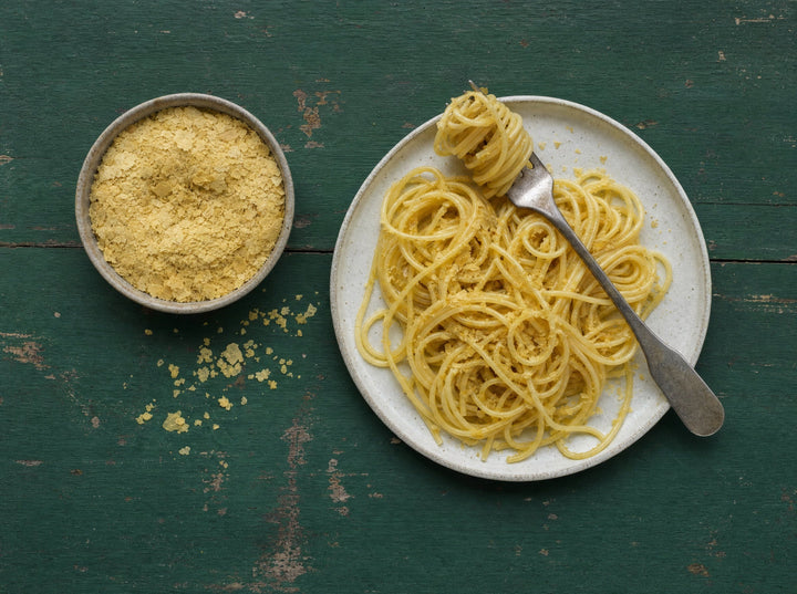 Eine Schale Nährhefeflocken neben einem Teller Spaghetti mit Nährhefe darauf
