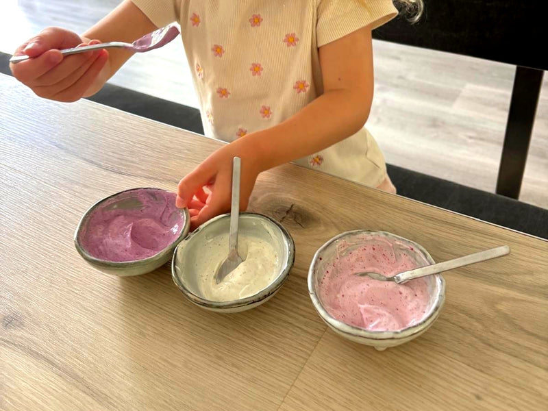 Drei Schälchen Joghurt in Pink, Weiß und Rosa auf einem Esstisch, Kinderhände mit Löffel
